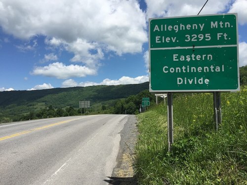 Sign indicating Allegheny Mountain elevation at 3295 feet and the Eastern Continental Divide, with a scenic background.