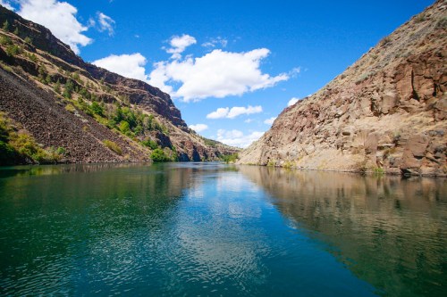 A serene river flows between rocky cliffs under a bright blue sky with fluffy clouds.