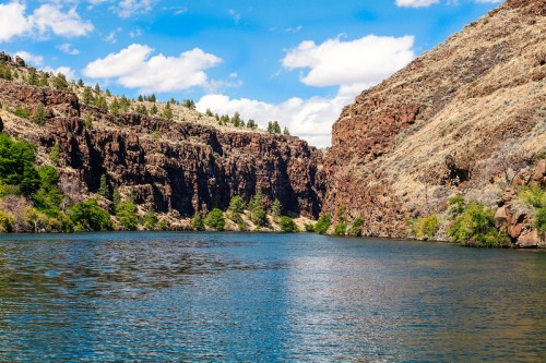 A serene river flows between rocky cliffs, surrounded by greenery under a bright blue sky with fluffy clouds.