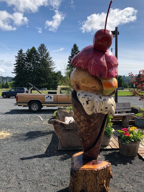 A large wooden sculpture of an ice cream cone with a cherry on top, set against a blue sky and trees.