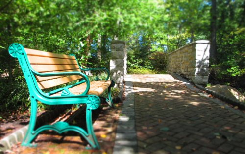 A green park bench beside a stone bridge, surrounded by lush greenery and a paved pathway.
