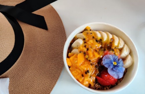 A bowl of fruit topped with banana, mango, passion fruit, and a flower, next to a wide-brimmed straw hat.