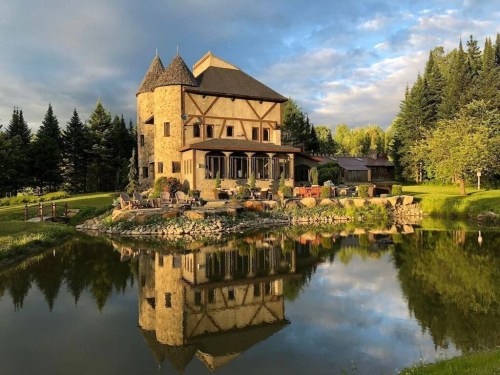 A picturesque stone house with a tower, reflecting in a calm pond, surrounded by lush greenery and trees.