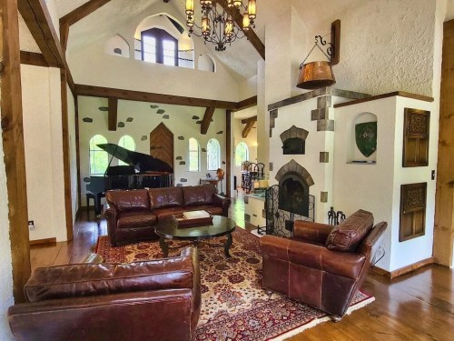 Cozy living room with leather sofas, a grand piano, and a rustic fireplace, featuring wooden beams and large windows.