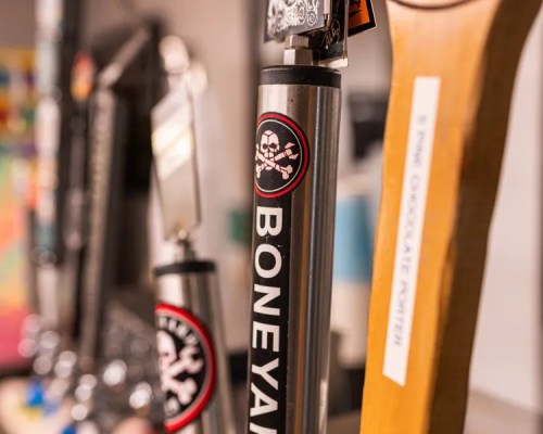 Close-up of beer taps featuring a "Boneyard" tap handle and others, with a blurred background of colorful bar decor.