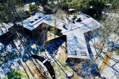 Aerial view of a modern house surrounded by trees and snow, featuring a unique geometric design and outdoor spaces.