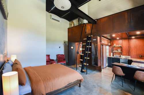 Cozy modern bedroom with a brown bedspread, kitchenette, and a lofted area, featuring industrial-style decor.