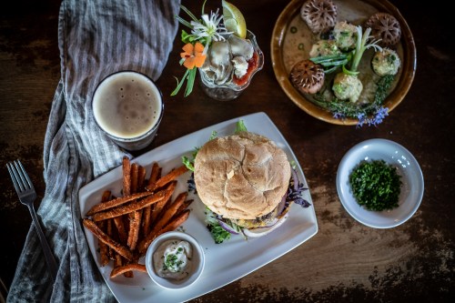 A delicious burger with sweet potato fries, a drink, and a plate of appetizers, all beautifully presented on a wooden table.