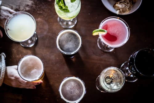 A variety of colorful cocktails and drinks arranged on a wooden table, with a hand reaching for one.