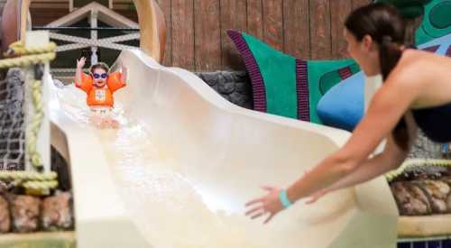 A child in an orange life jacket slides down a water slide, while an adult reaches out to catch them.