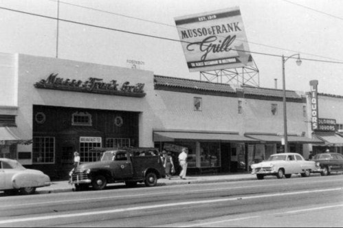 Black and white photo of Musso & Frank Grill with vintage cars parked outside and a liquor store nearby.