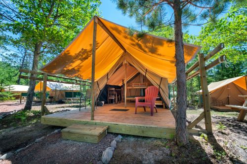 A cozy glamping tent with an orange canopy, wooden deck, and a red chair, surrounded by trees in a sunny outdoor setting.
