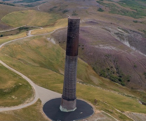 A tall, cylindrical structure stands alone on a grassy landscape, surrounded by rolling hills and a winding road.