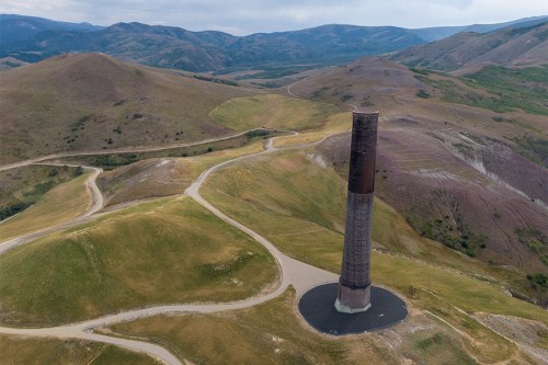 A tall, cylindrical structure stands on a grassy hillside, surrounded by rolling mountains and winding dirt roads.