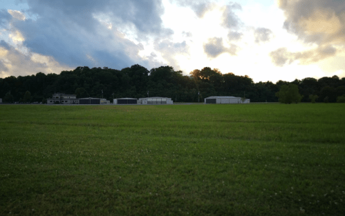 A grassy field with storage buildings in the background, under a cloudy sky at sunset.