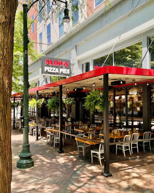 Outdoor seating area of Aldo's Pizza Pies, featuring wooden tables, chairs, and hanging plants under a red awning.