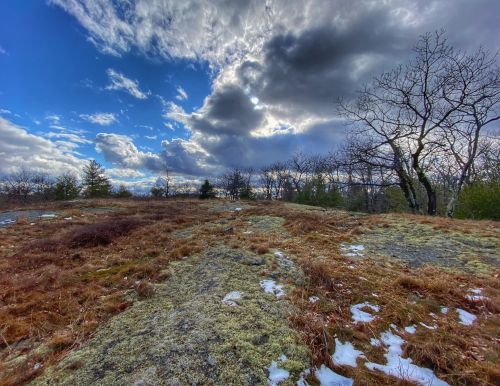A scenic landscape featuring rocky terrain, sparse trees, and a dramatic sky with clouds and sunlight peeking through.
