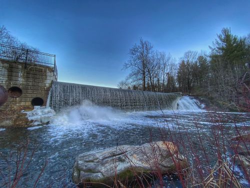 A serene waterfall cascades over a dam, surrounded by trees and a clear blue sky.