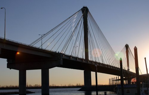 Silhouette of a cable-stayed bridge at sunset, with water and a dock in the foreground.