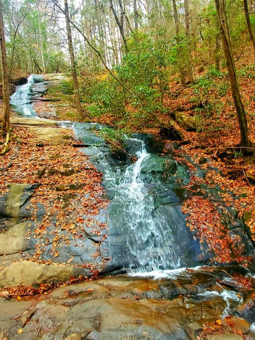A serene waterfall cascades over rocky terrain, surrounded by trees and autumn leaves.