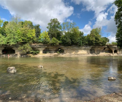 A serene river scene with rocky banks, lush greenery, and a cloudy blue sky reflecting on the water's surface.
