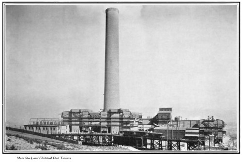 Black and white image of a large industrial building with a tall smokestack, set on a raised platform.