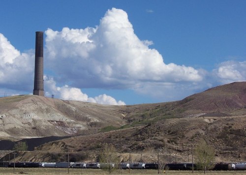 A tall smokestack rises above a barren landscape with rolling hills and a train of freight cars in the foreground.