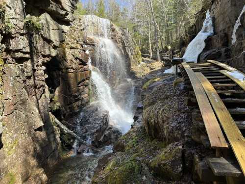 A scenic view of a waterfall cascading down rocky cliffs, with a wooden path alongside a flowing stream.