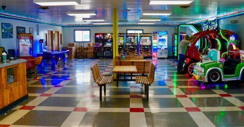 A colorful arcade room with games, vending machines, and benches, featuring a vintage car ride and pool table.