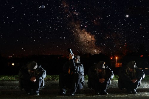 A person in a dark coat squats under a starry sky, gazing at the Milky Way with a telescope.