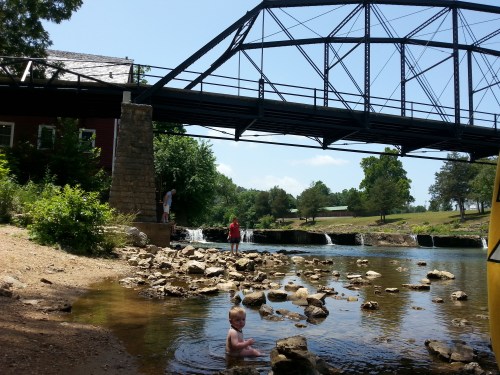 A child wades in shallow water by a rocky shore, with a bridge overhead and people in the background near a waterfall.