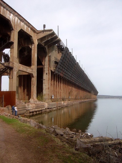 A large, weathered industrial structure extends over a calm body of water, with rocky shoreline and a path nearby.