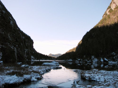 A serene winter landscape featuring a frozen river surrounded by snow-covered mountains and trees under a clear sky.