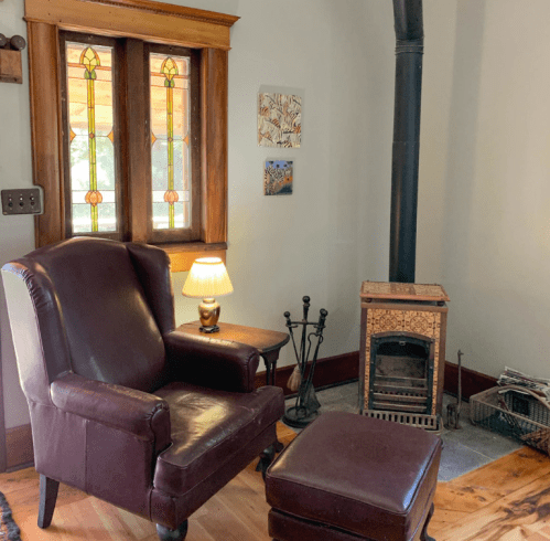 Cozy living room corner with a leather armchair, lamp, and a small wood stove beside a stained glass window.