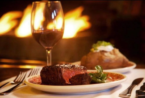 A plate with a juicy steak, a baked potato, and a glass of red wine, set against a warm, glowing fireplace.