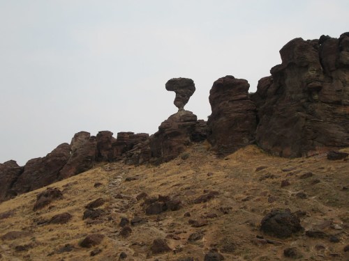 A unique rock formation resembling a figure, perched atop a rocky hillside with a cloudy sky in the background.
