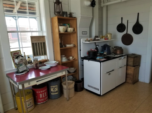 A vintage kitchen with a white stove, wooden shelves, dishes, and various kitchenware on display.
