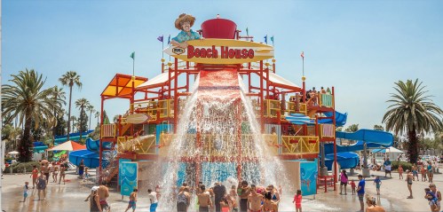 A colorful water play structure with slides and a large splash feature, surrounded by palm trees and people enjoying the sun.