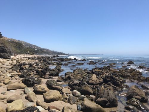 A rocky beach with clear blue skies, gentle waves, and distant hills under bright sunlight.