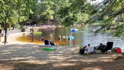 A sunny lakeside scene with people swimming, lounging on the shore, and enjoying inflatable floats in the water.