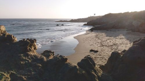 A serene beach scene with rocky formations, gentle waves, and a distant figure walking along the shore at sunset.