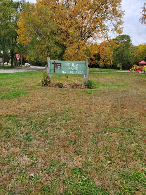 Sign for Beulah Park Nature Area surrounded by grass and trees with autumn foliage. Playground visible in the background.