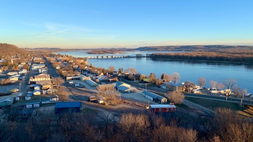 A scenic view of a river town with buildings, trees, and a bridge over the water, under a clear blue sky.