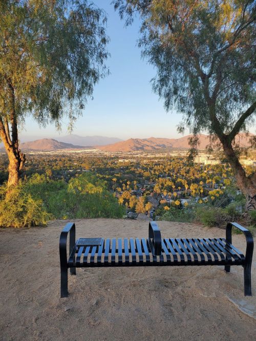 A black bench overlooks a scenic view of mountains and a valley, framed by trees under a clear sky.
