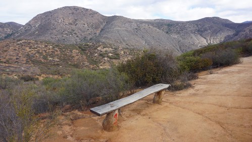 A wooden bench sits on a dirt path, surrounded by hills and sparse vegetation under a cloudy sky.
