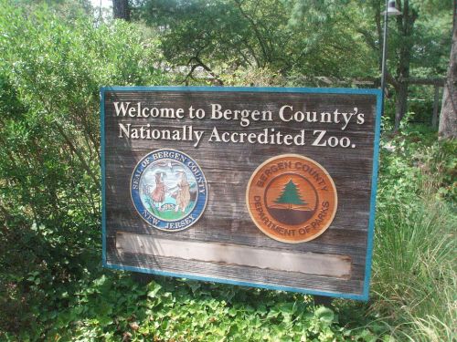 Sign welcoming visitors to Bergen County's nationally accredited zoo, featuring county seals and surrounded by greenery.