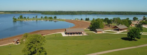 A scenic view of a lake with sandy shores, green trees, and picnic pavilions under a clear blue sky.