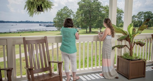 Two women stand on a balcony overlooking a river, enjoying drinks and the scenic view on a sunny day.