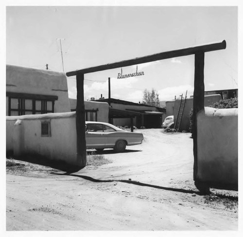 Black and white photo of a rustic entrance with a sign reading "Blumenschein" and a car parked nearby.