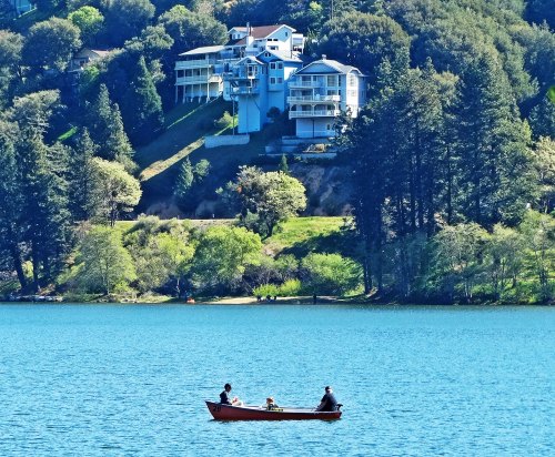 A small boat with three people floats on a blue lake, surrounded by lush greenery and hillside homes.
